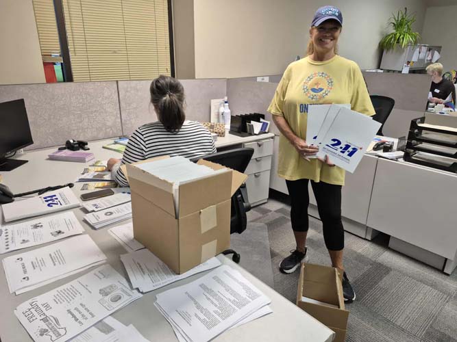A volunteer holding 2-1-1 signs beside boxes of printed materials in an office