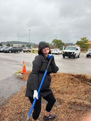 A volunteer resting on a shovel beside a mulch pile in a parking lot