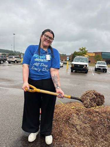A volunteer with a shovel beside a mulch pile in a parking lot