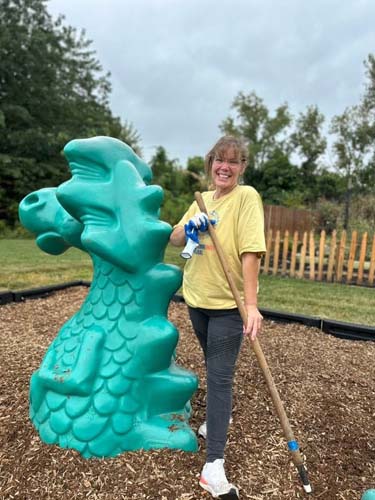 A volunteer raking mulch beside a teal dragon playground sculpture