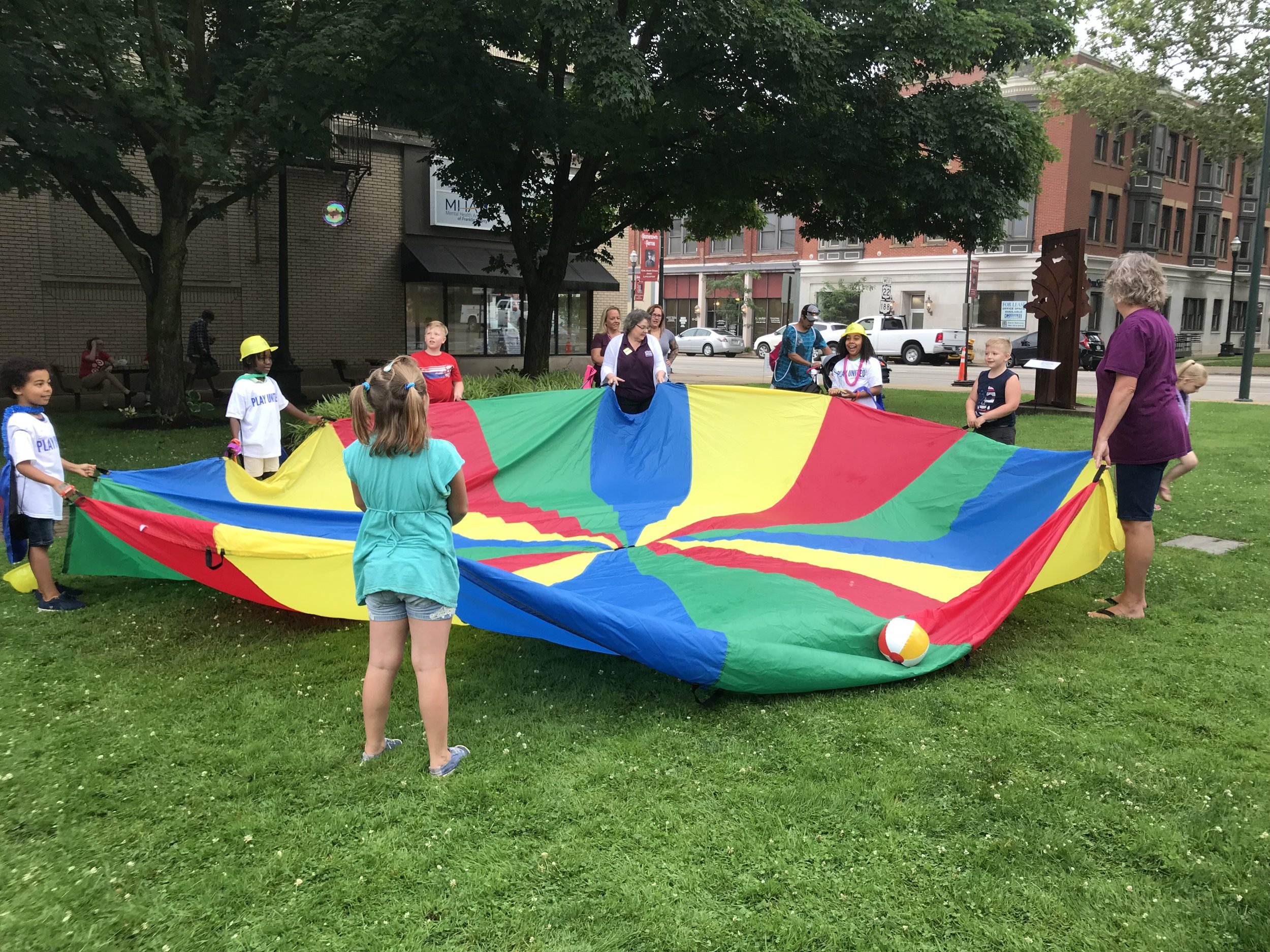 Children and volunteers playing with a rainbow parachute on the lawn downtown
