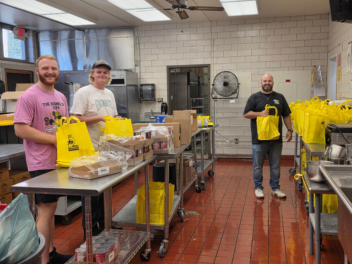 Three volunteers smiling while packing yellow Full Plate Club bags at stainless steel tables in a school kitchen